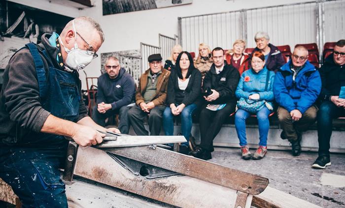 a craftsman in blue overalls sits on a bench working the piece of slate with a saw. in the background, a group of visitors are watching him. He explai